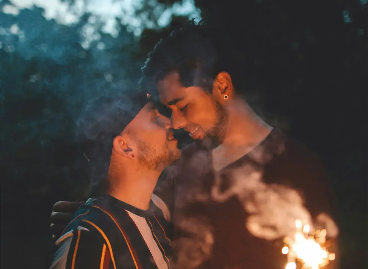 Coppia gay che si bacia con scintille e fumo, un momento intimo e passionale.