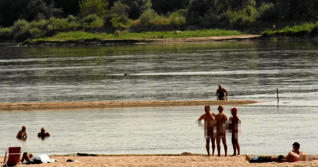 Ludzie opalają się i kąpią na plaży nudystów. Woda jest spokojna, a na brzegu widać kilka osób.