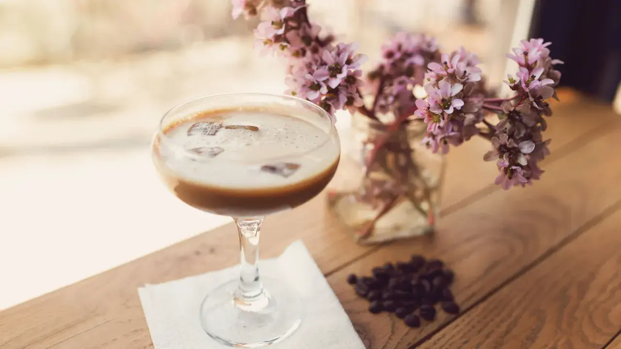 Ein cremiger Eiskaffee-Cocktail mit Eiswürfeln, daneben zarte rosa Blüten und Kaffeebohnen auf Holztisch.