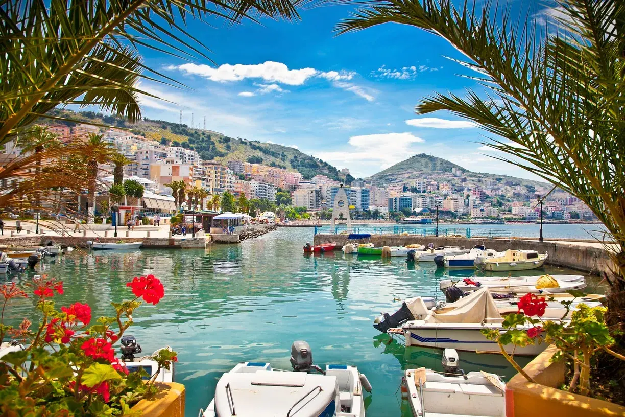 Albania, gdzie najpiękniej! Widok na port z palmami, kolorowymi budynkami na wzgórzu i łodziami na turkusowej wodzie.