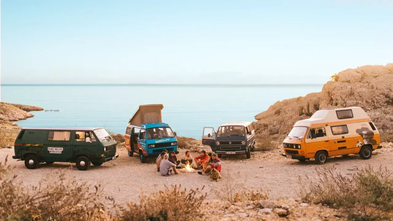 Vier bunte Campervans am Meer, Menschen sitzen am Lagerfeuer.