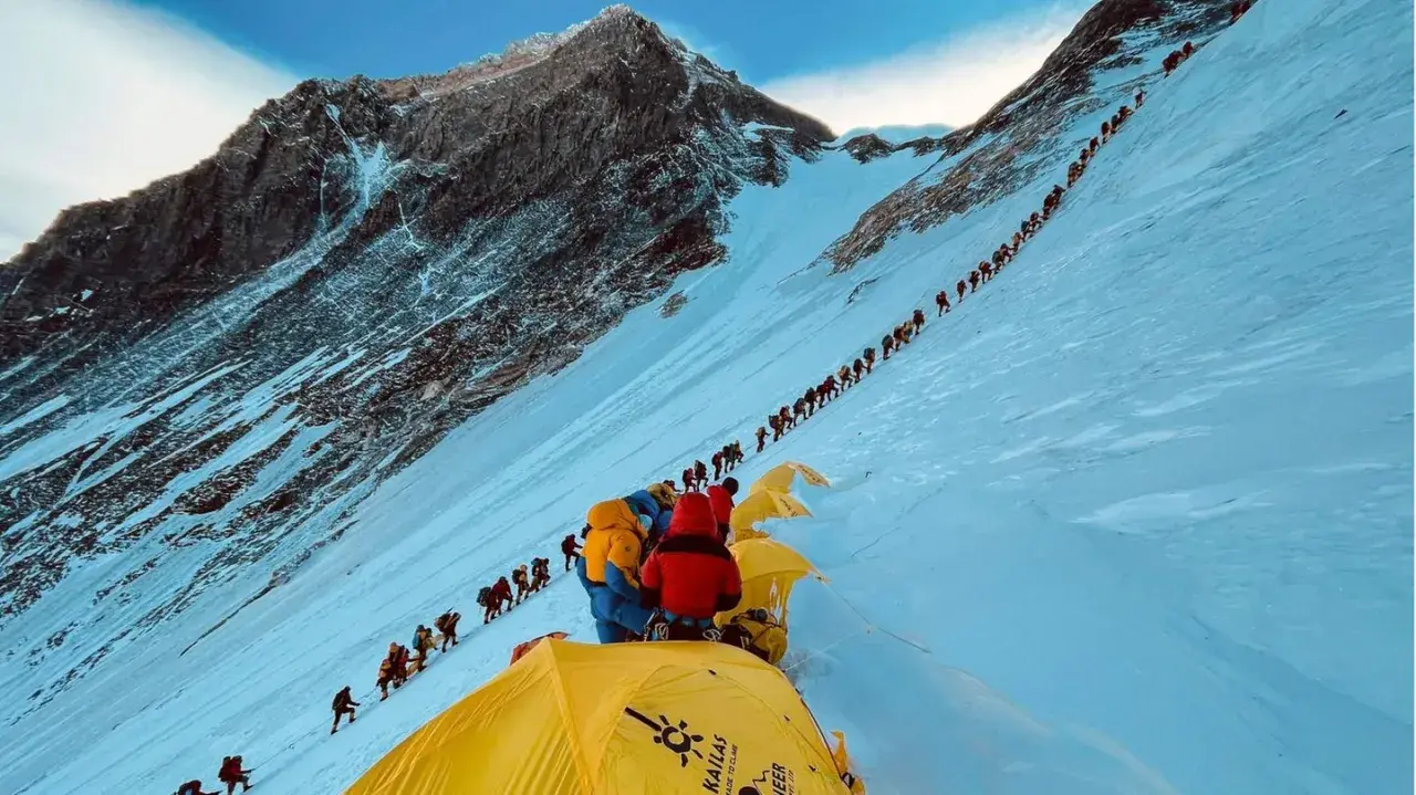 Eine lange Schlange von Bergsteigern kämpft sich den schneebedeckten Hang des Mount Everest hinauf. Die Frage, wie lange dauert es den Mount Everest zu besteigen, wird hier durch die schiere Anzahl der Menschen beantwortet.