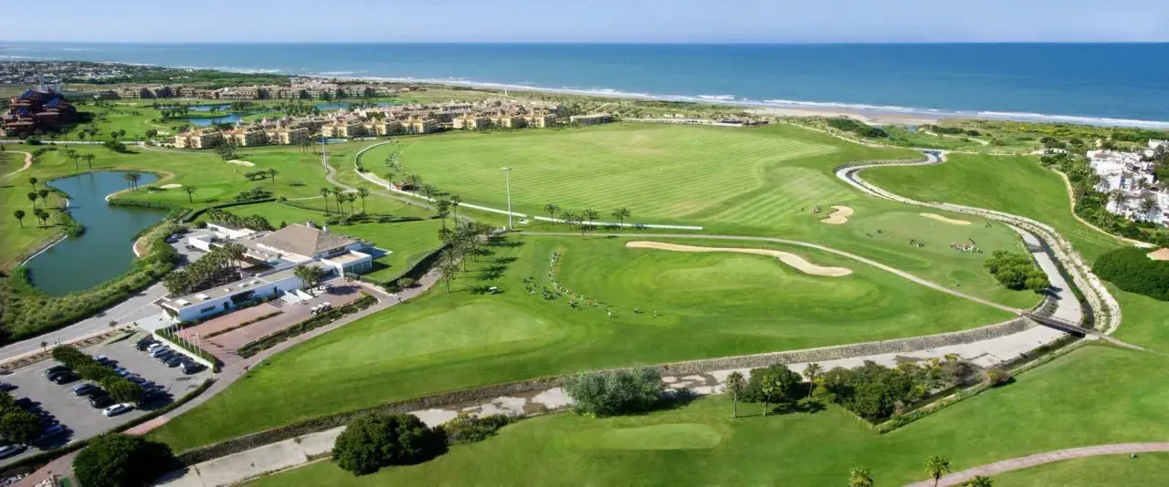 Vista aérea del campo de golf del hotel Barceló Costa Ballena, con el mar azul al fondo y edificios residenciales.