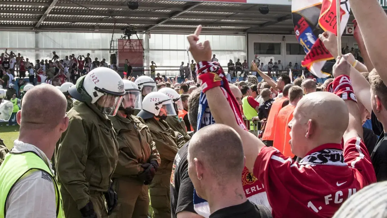 Policja w pełnym rynsztunku stoi naprzeciwko grupy kibiców, wśród których widać ultrasi z flagami i szalikami.
