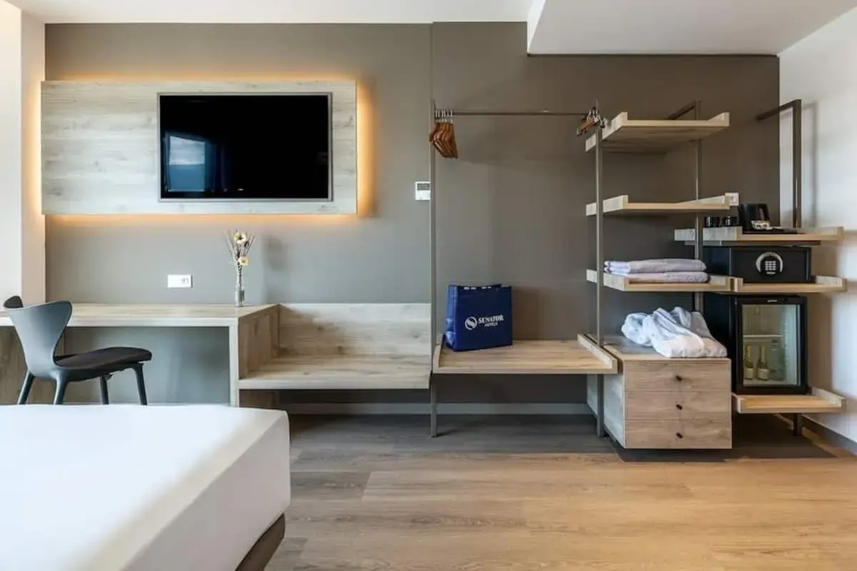Habitación moderna con TV, escritorio, armario abierto con minibar y caja fuerte. Una bolsa azul con el logo de Senator Hotels descansa sobre un banco.