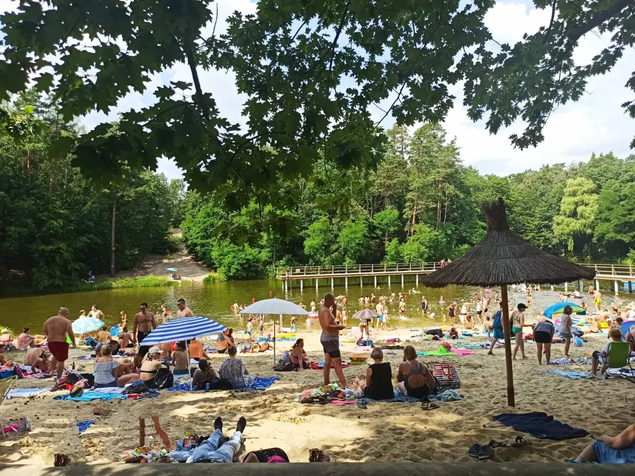 Słoneczny dzień na plaży przy zalewie w Garbatce. Ludzie odpoczywają na piasku, kąpią się w wodzie i spacerują po pomoście.