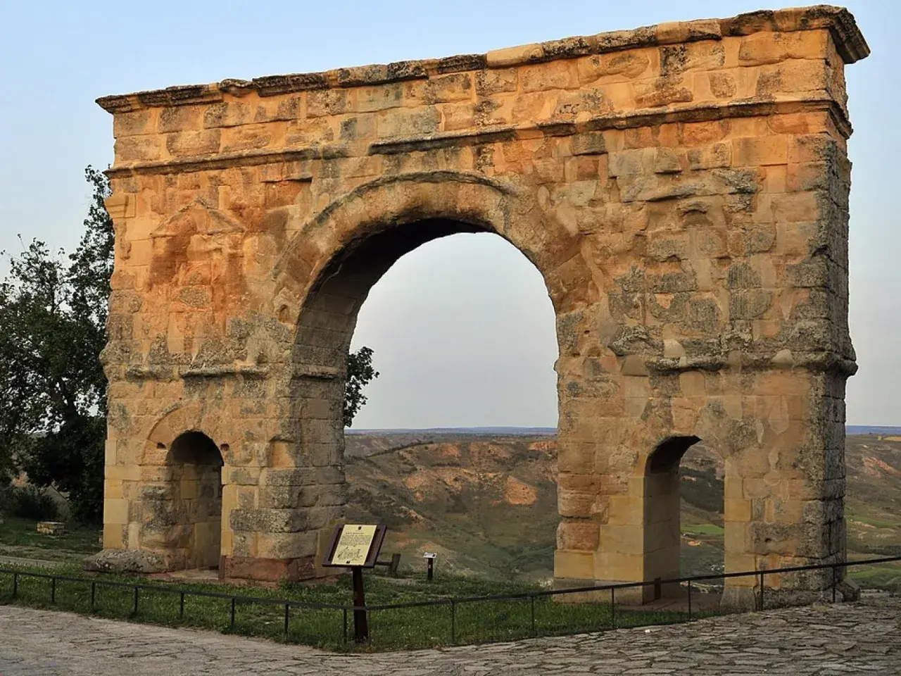 Imponente arco romano de piedra, con dos arcos menores a los lados, se alza sobre un paisaje de colinas verdes.