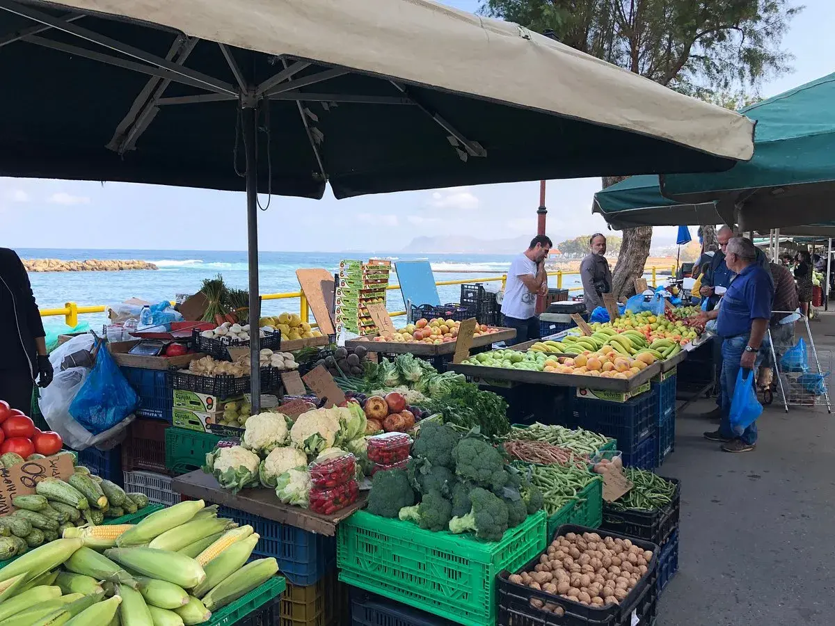 Frukt- och grönsaksstånd vid marknad i Chania. Färska varor som majs, broccoli och tomater säljs med havet i bakgrunden.