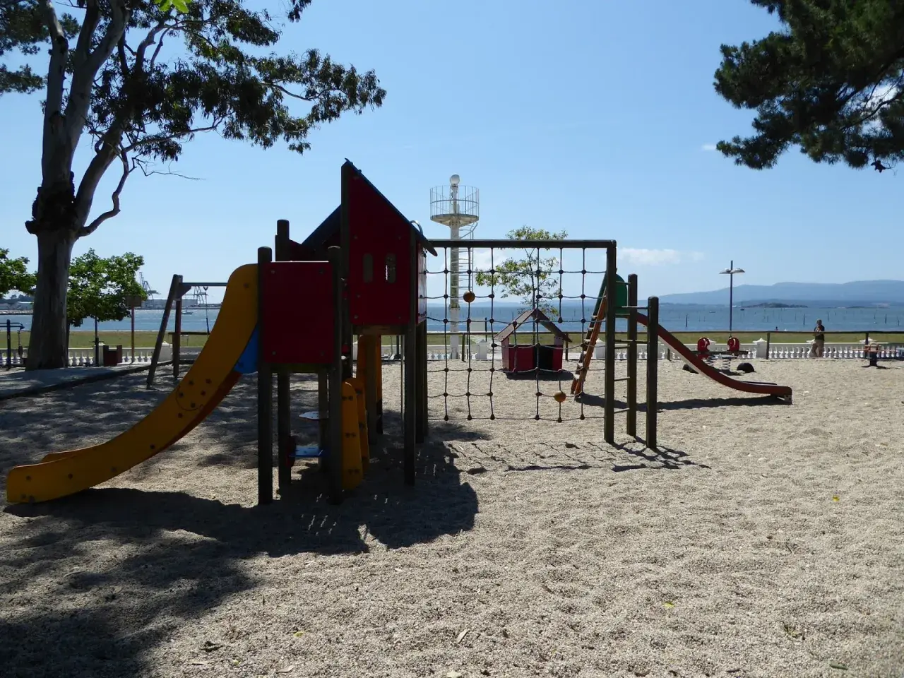 Un colorido parque infantil en la playa invita a jugar. Toboganes, redes y columpios esperan bajo el sol, con el mar de fondo.