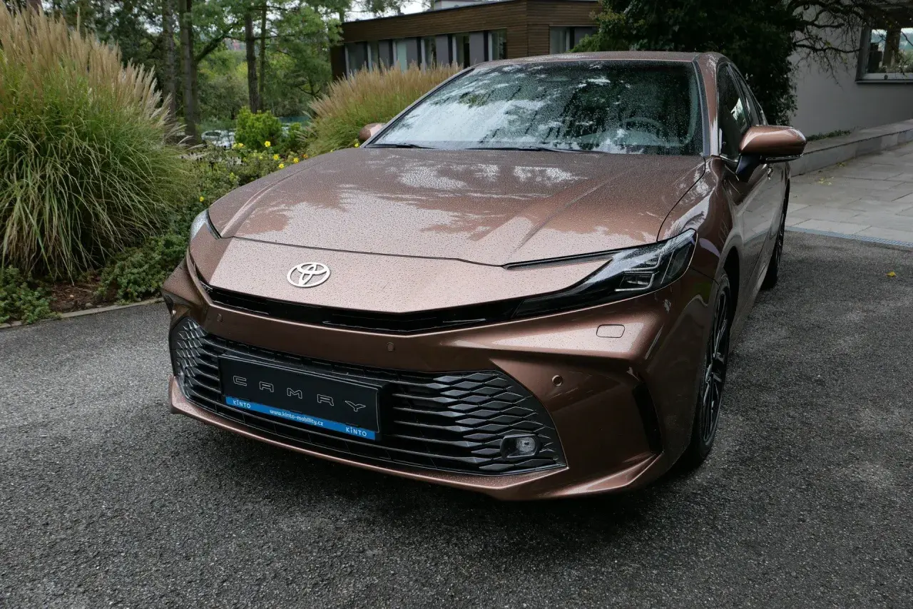 Nowa Toyota Camry 2024 w kolorze brązowym, z kroplami deszczu na karoserii, zaparkowana przed nowoczesnym budynkiem.