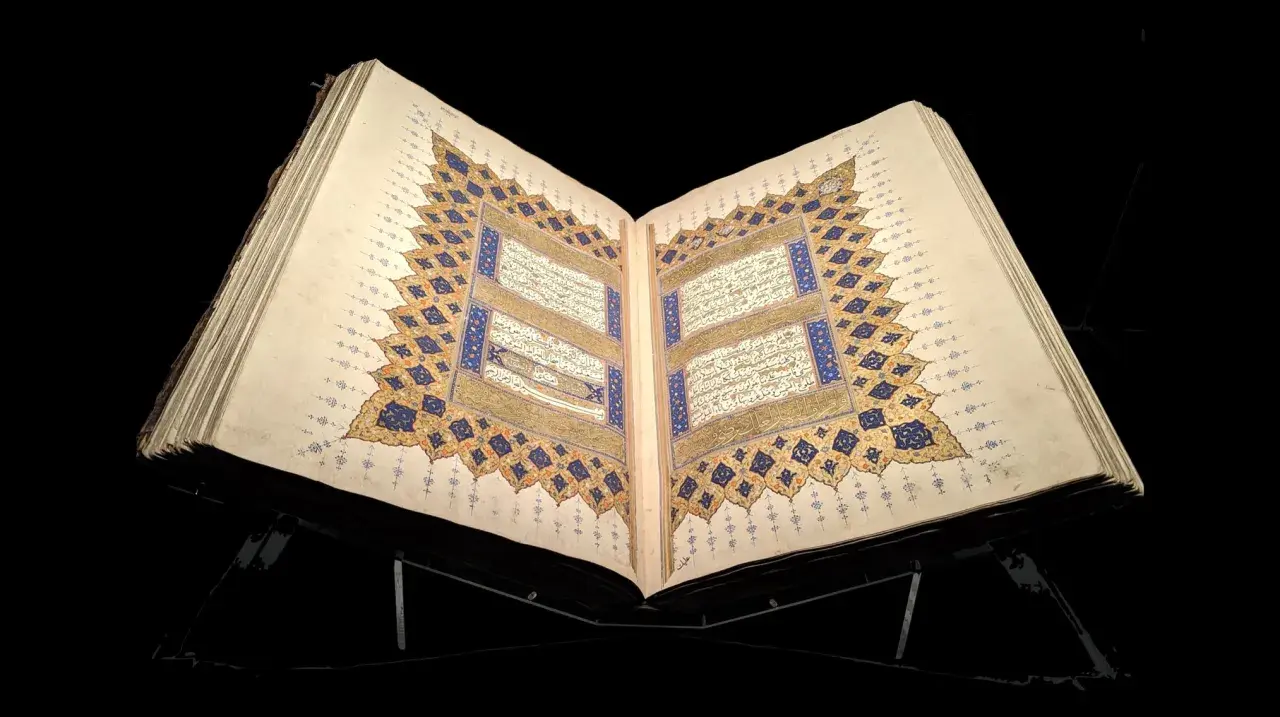 En öppen, gammal Koran på ett ställ, med detaljerad kalligrafi och gyllene ornament. En påminnelse om vem som skrev Koranen.