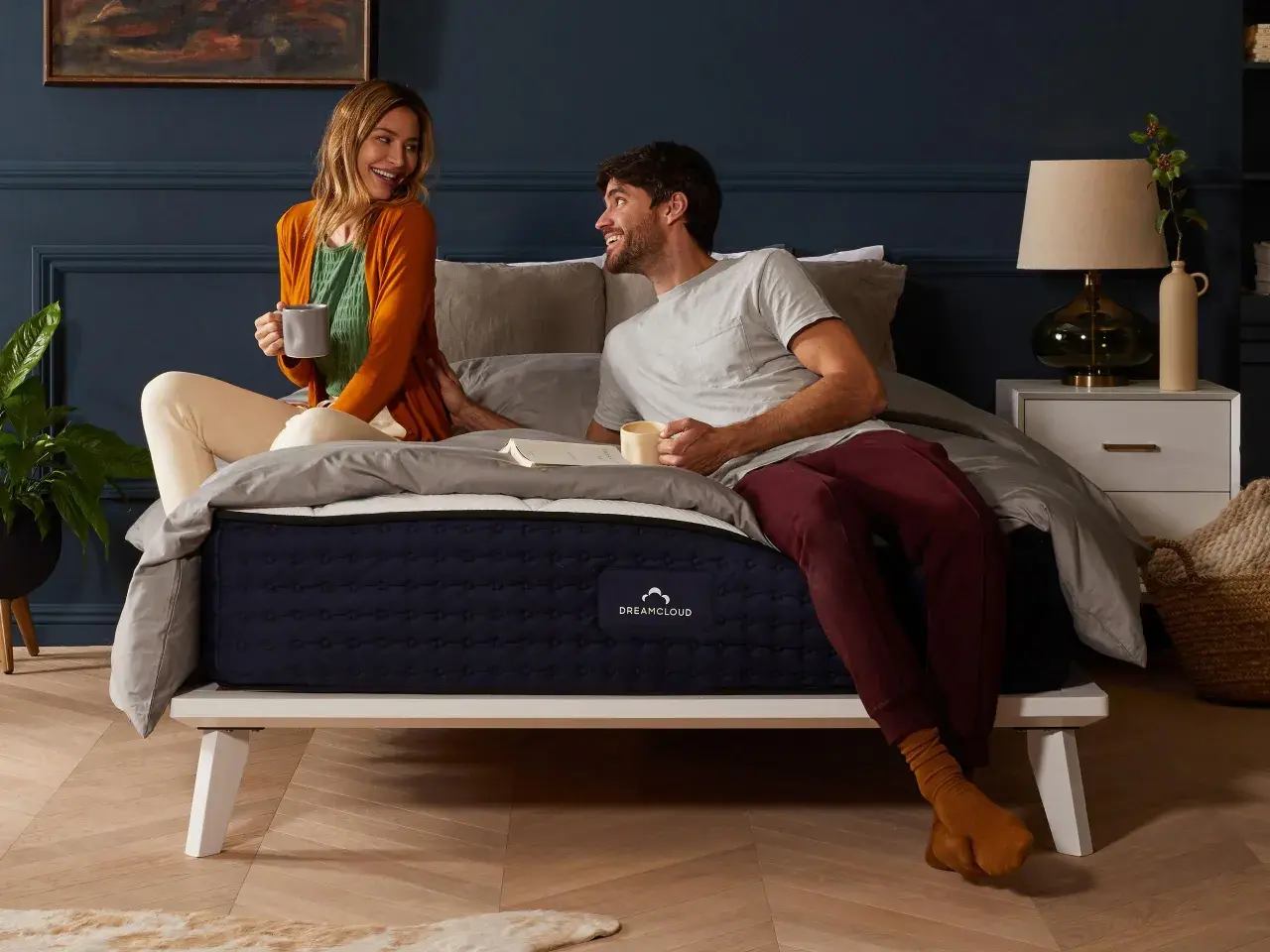 A couple enjoys coffee in bed. The DreamCloud hybrid mattress is on a white platform bed, answering the question: can you put a hybrid mattress on a box spring?