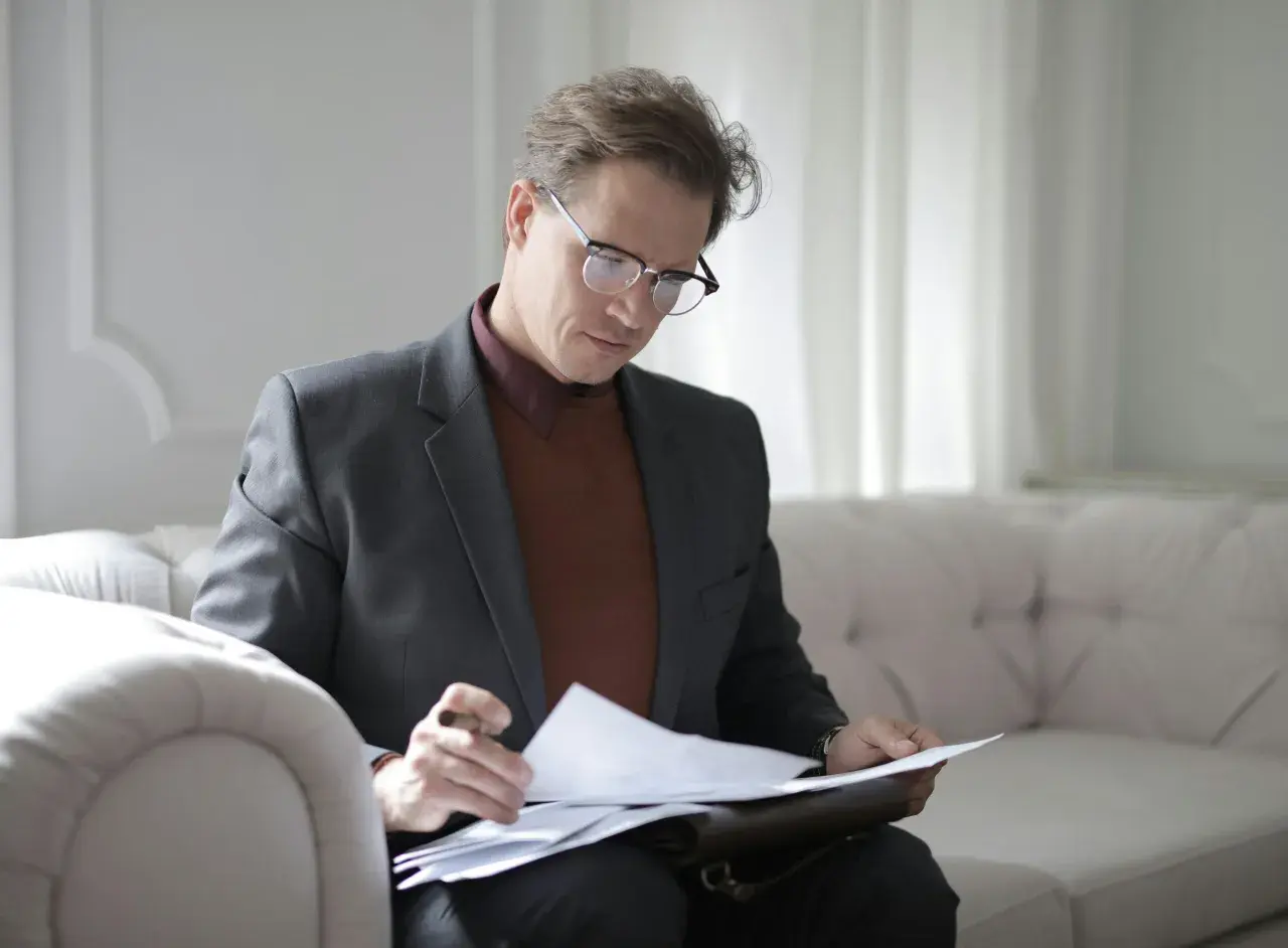 Hombre con gafas revisa documentos en un sofá, concentrado en su trabajo. ¿Qué es la fein? Es la dedicación a los detalles.