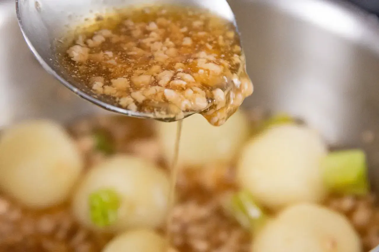 A spoon pours a thick, savory ankake sauce over tender dumplings and vegetables in a pot.