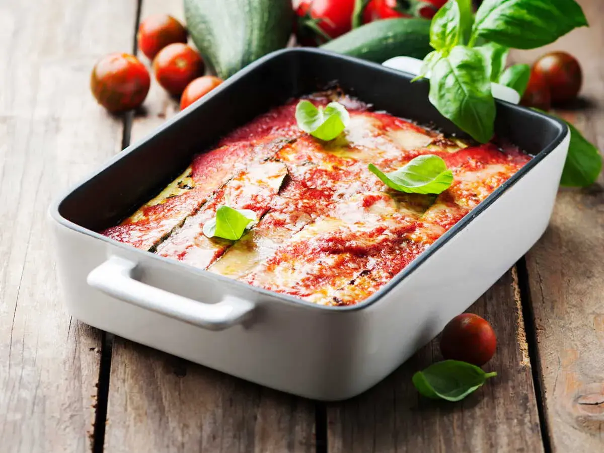 Lasagne courgette gratinée, garnie de basilic frais, sur fond de bois rustique avec des tomates cerises et des courgettes.