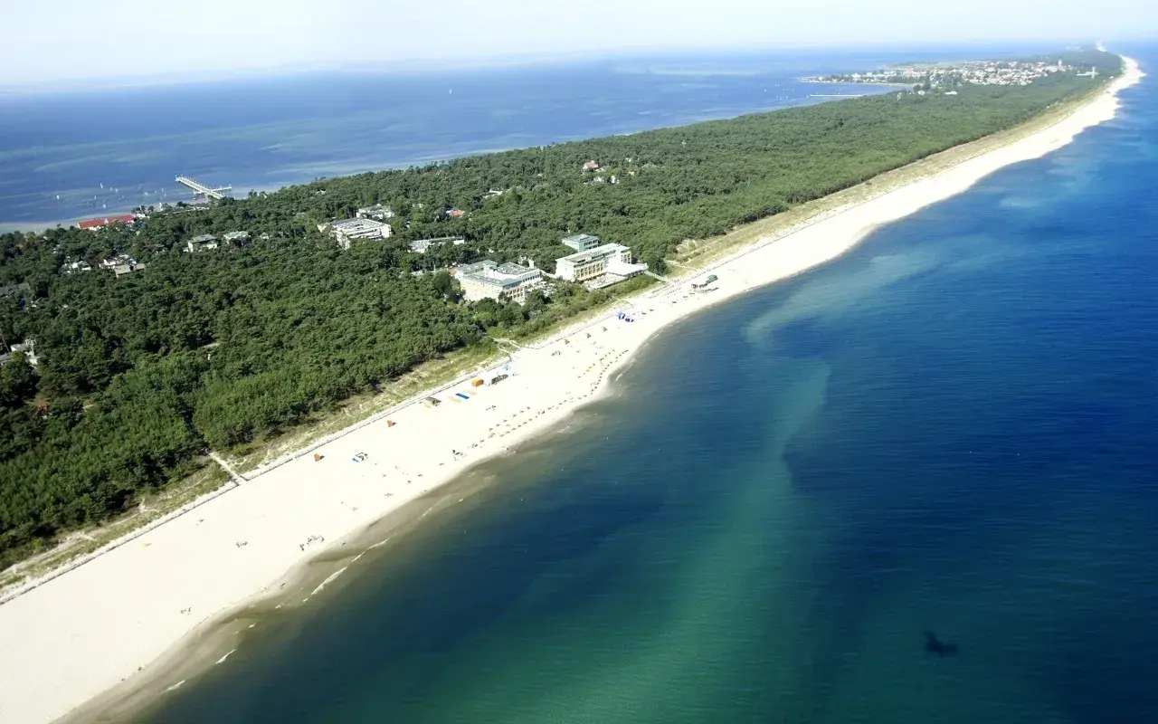 Jurata plaża z lotu ptaka: szeroka, piaszczysta plaża, sosnowy las i budynki hotelowe nad turkusowym morzem.