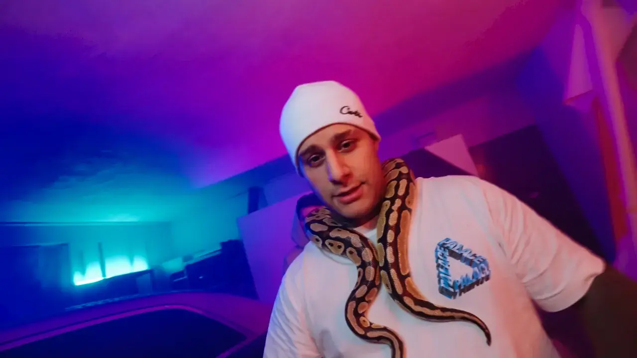 Macias w białej czapce i t-shircie z wężem na szyi, w neonowym świetle.
