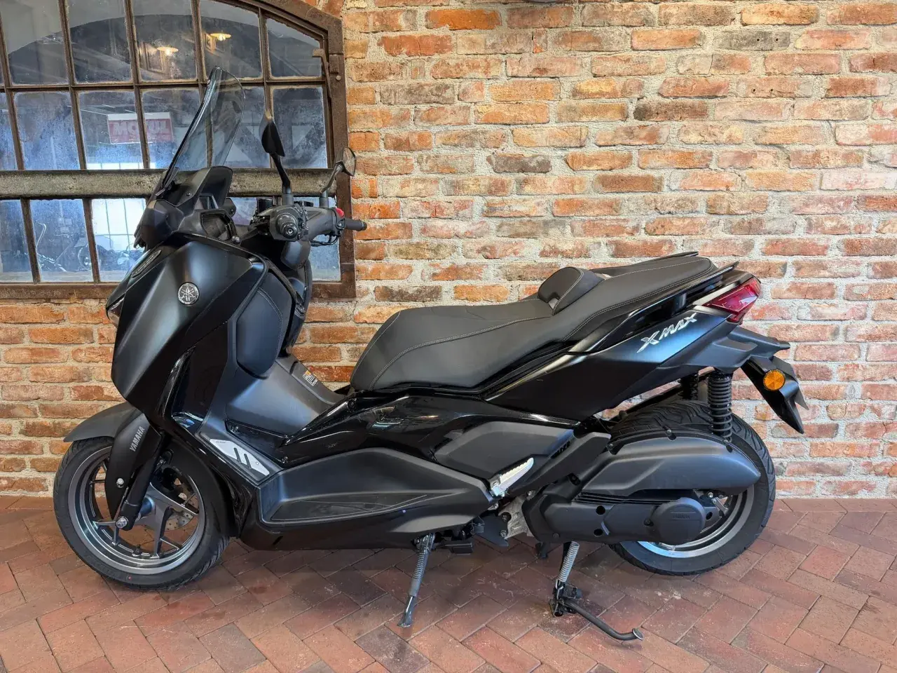 Czarny skuter Yamaha X-Max 125 na tle ceglanej ściany i okna.