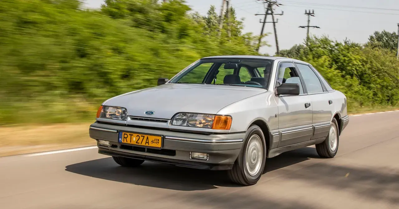 Srebrny Ford Scorpio jedzie po drodze, otoczony zielenią.