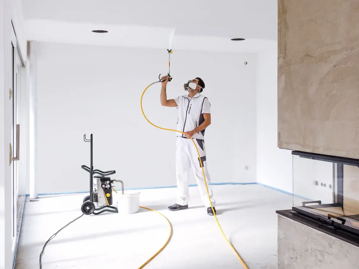 Pittore con maschera protettiva usa una pistola per vernice a spruzzo per dipingere il soffitto di una stanza vuota.