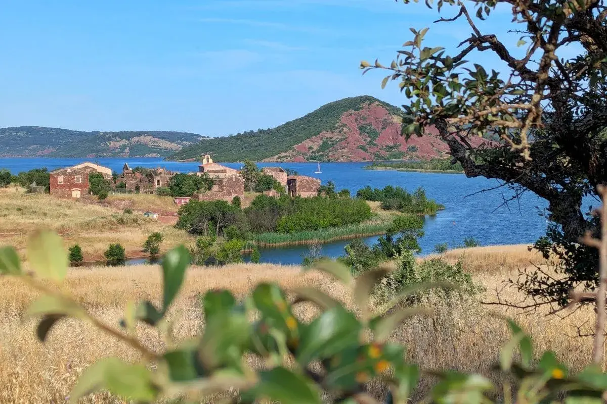 Vue sur le lac du Salagou, avec ses eaux bleues, ses collines rouges et les ruines d'un village. Un voilier navigue paisiblement.