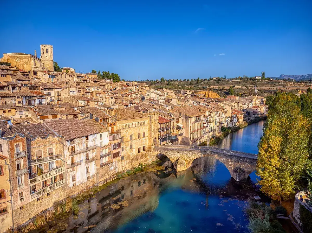 Pueblo con casas de tejas de barro y un puente de piedra sobre un río azul, evocando la toscana española.