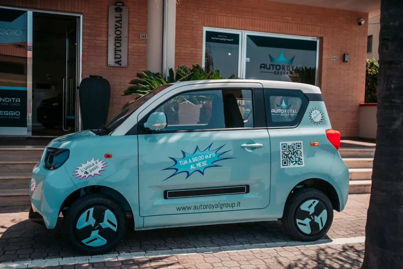 Macchina piccola 4 posti azzurra, con adesivi promozionali "TUA A 99,00 euro AL MESE" e QR code.