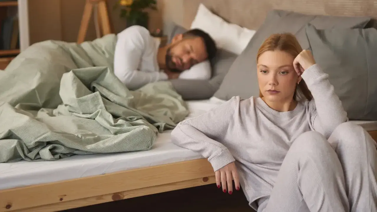 Mujer triste sentada en el suelo, mientras su pareja duerme en la cama. Reflexiona sobre qué significa soñar que tu pareja te es infiel.