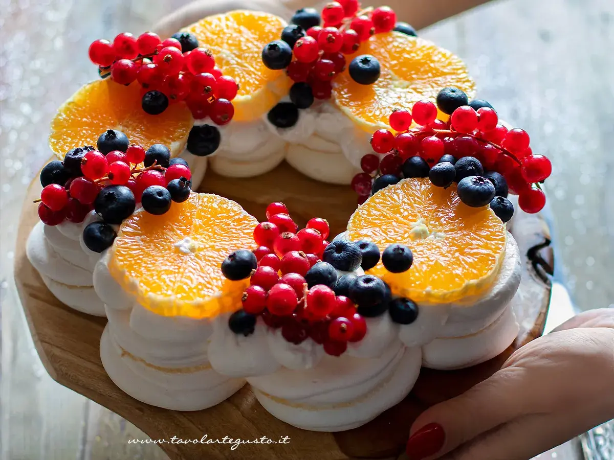 Torta pavlova a forma di corona, decorata con fette d'arancia, ribes rossi e mirtilli.