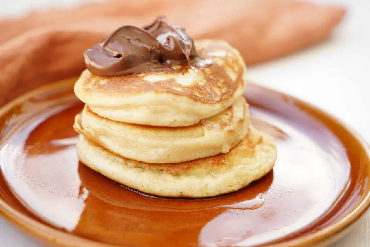 En hög med fluffiga amerikanska pannkakor toppas med chokladkräm på en brun tallrik.