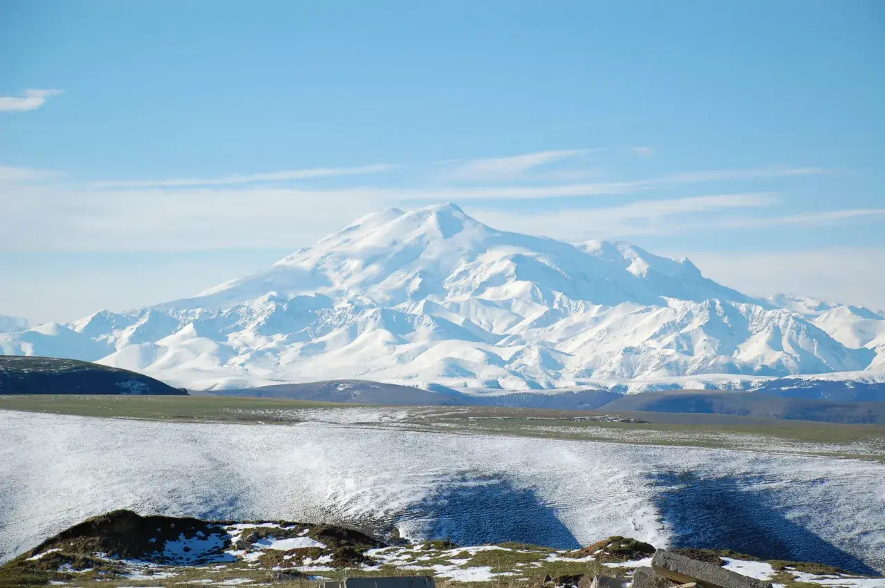 Majestatyczny, ośnieżony najwyższy szczyt Kaukazu, Elbrus, góruje nad pokrytymi śniegiem wzgórzami i zielonymi dolinami.