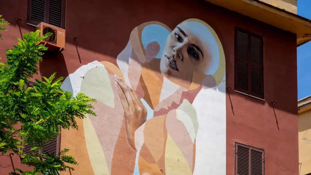 Un volto intenso e mani giunte su un muro colorato in via di Tor Marancia, un'opera d'arte che cattura lo sguardo.