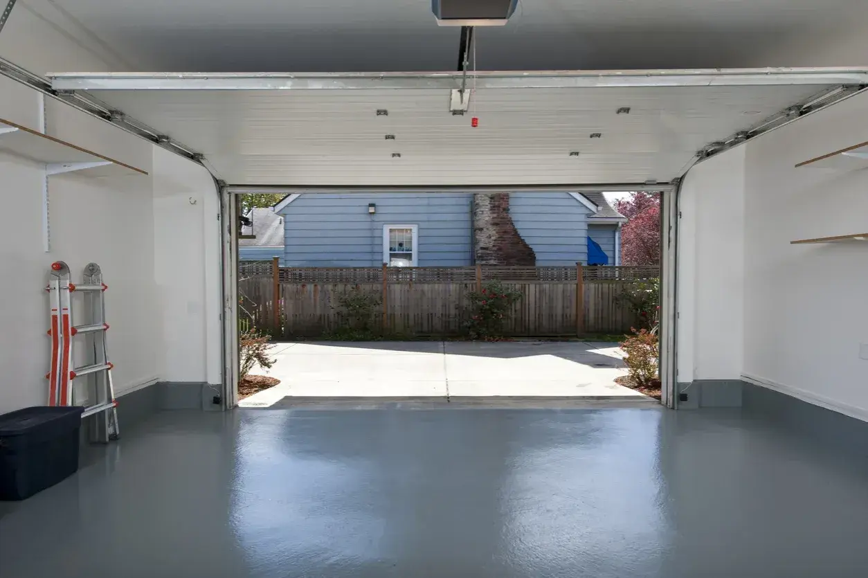 Interno di un garage con porta sezionale aperta su un cortile. Si vede una scala e un contenitore. Pensare a quanto costa trasformare un garage in abitazione.