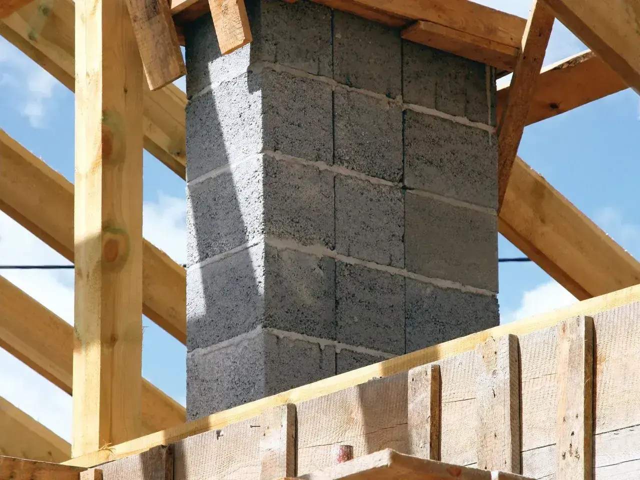 Budowa komina systemowego z bloczków betonowych, widoczny fundament i drewniana konstrukcja dachu.