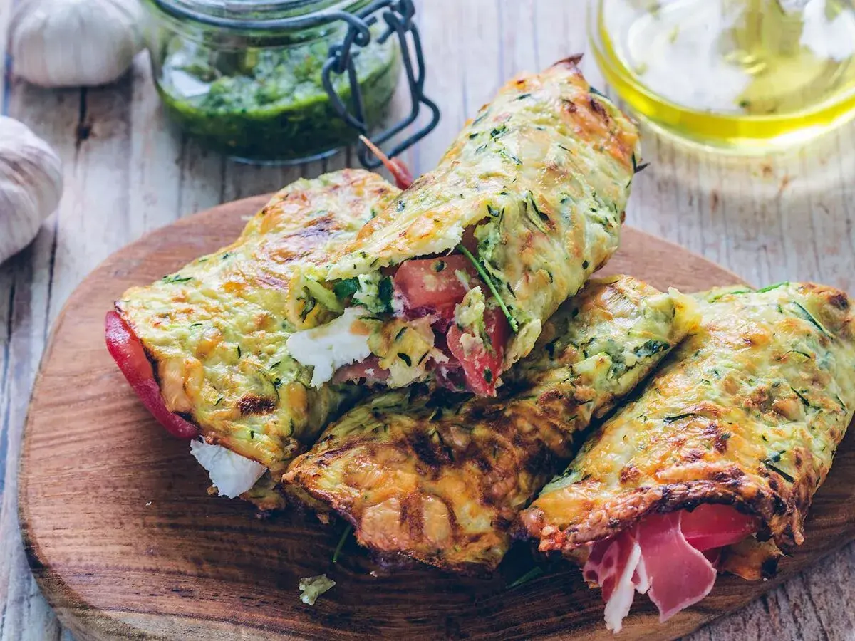 Złociste tortilla z cukinii, nadziewane serem, pomidorami i szynką, prezentowane na drewnianej desce.