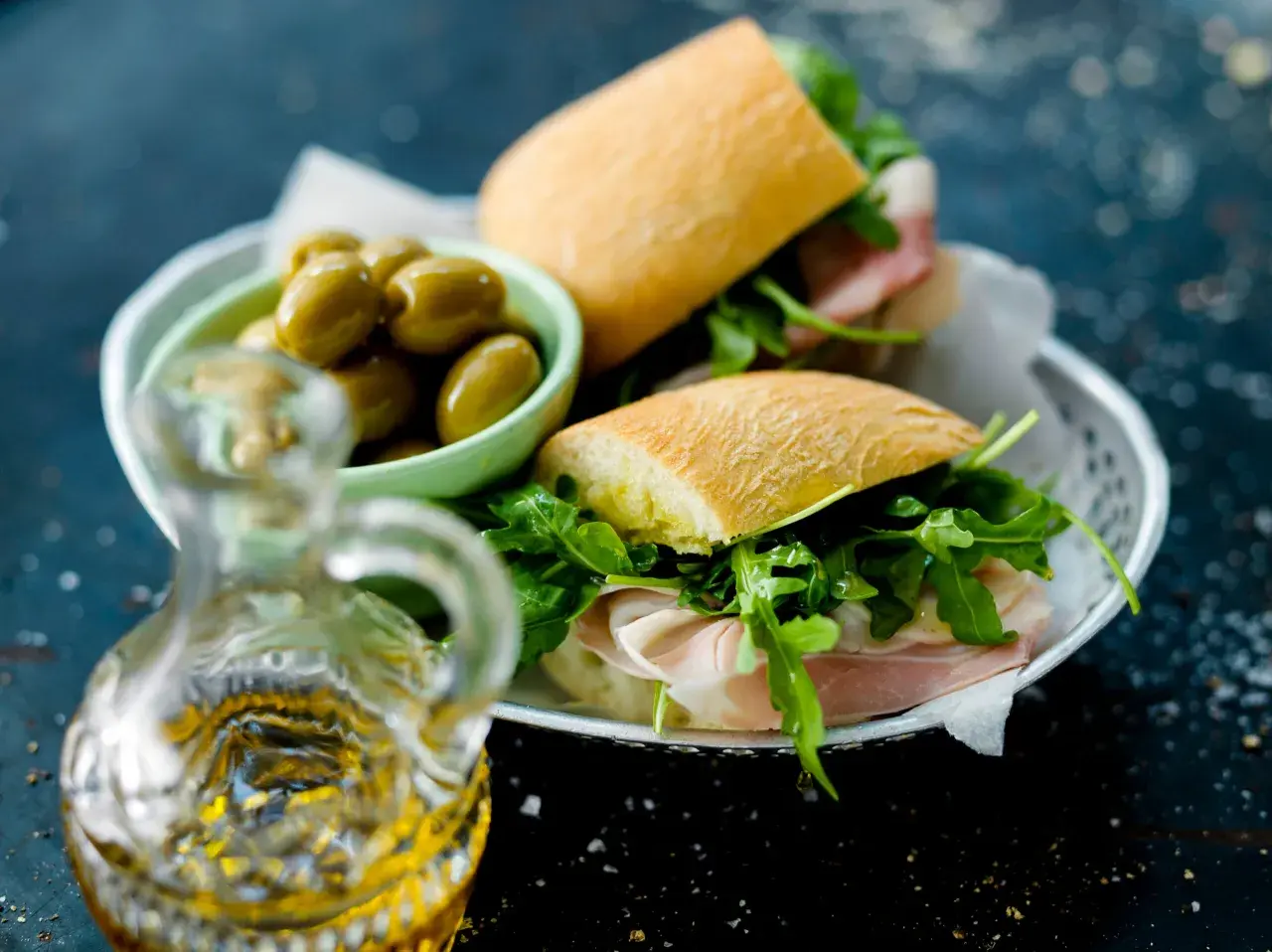 Två halvor av en ciabatta macka med prosciutto och ruccola, serverade med gröna oliver och en karaff olivolja.