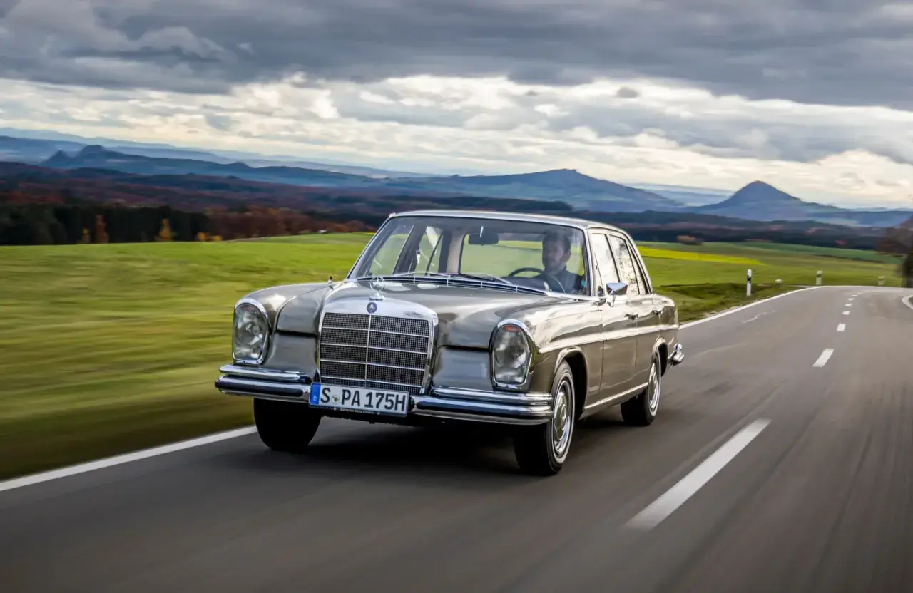 En klassisk Mercedes 60-tal glider fram på en slingrande landsväg med böljande gröna fält och dramatiska berg i bakgrunden.