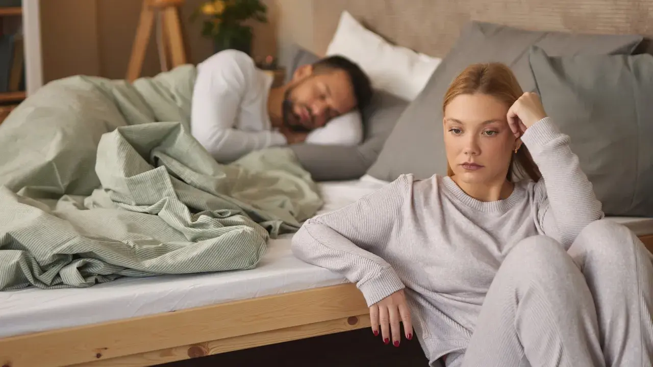 Eine Frau sitzt nachdenklich am Bettrand, während ihr Partner schläft. Sie kämpft mit ihrer Sexsucht bei Frauen.
