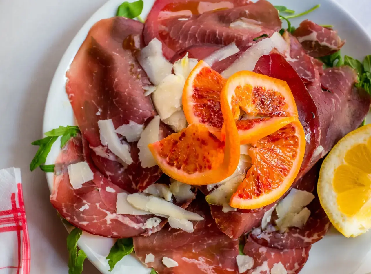 Wykwintna szynka bresaola z płatkami parmezanu, krwistymi pomarańczami i cytryną na talerzu.