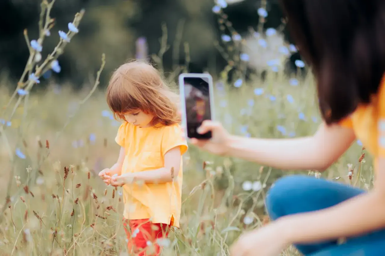 Dziecko w żółtej koszulce zbiera kwiatki na łące, a mama dokumentuje chwilę telefonem, praktykując sharenting.