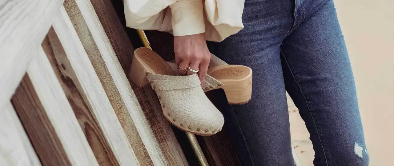 Une femme tient un sabot femme en toile beige, orné de clous, avec un talon en bois. Elle porte un jean bleu.