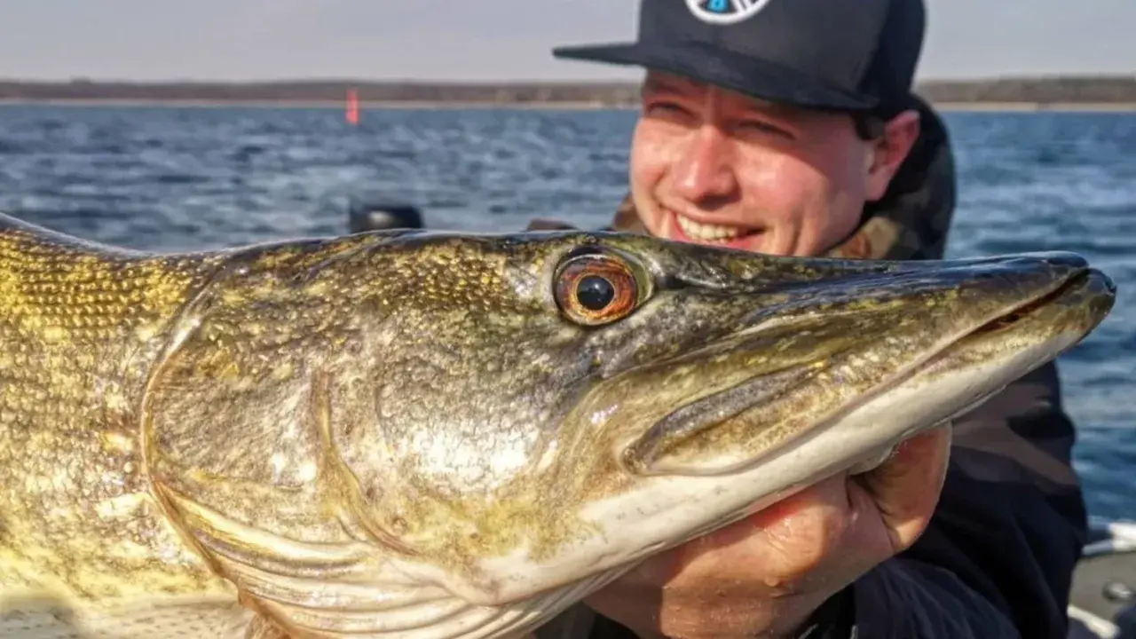 Glücklicher Angler hält stolz einen großen Hecht, gefangen nach einem Gewitter. Das Wasser ist ruhig, die Sonne scheint.