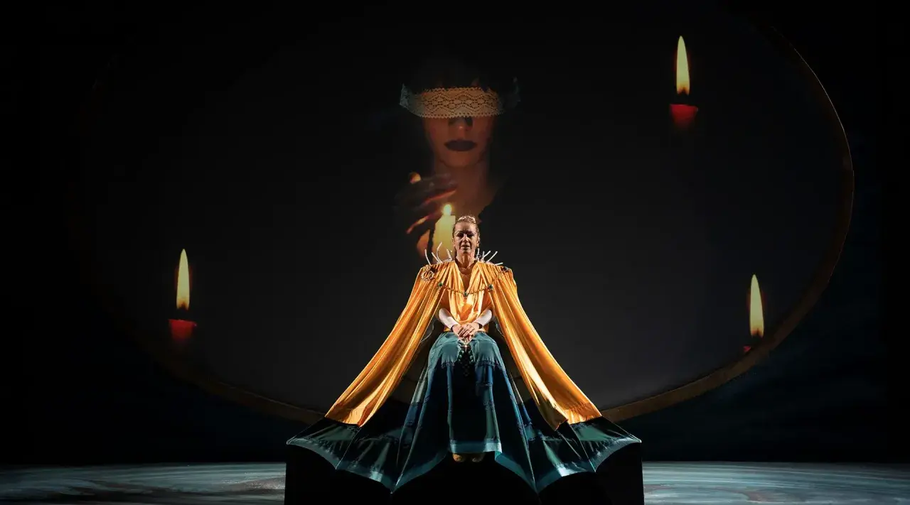 Scena teatrale ispirata al circe film: una donna con mantello dorato, circondata da candele, sotto un'immagine proiettata di una figura bendata.