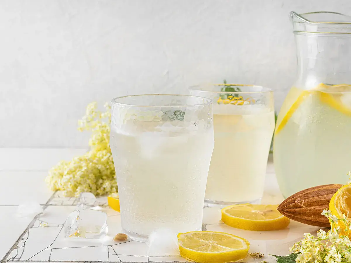 Deux verres de citronnade maison rafraîchissante avec des glaçons, des rondelles de citron, des fleurs de sureau et une carafe remplie.
