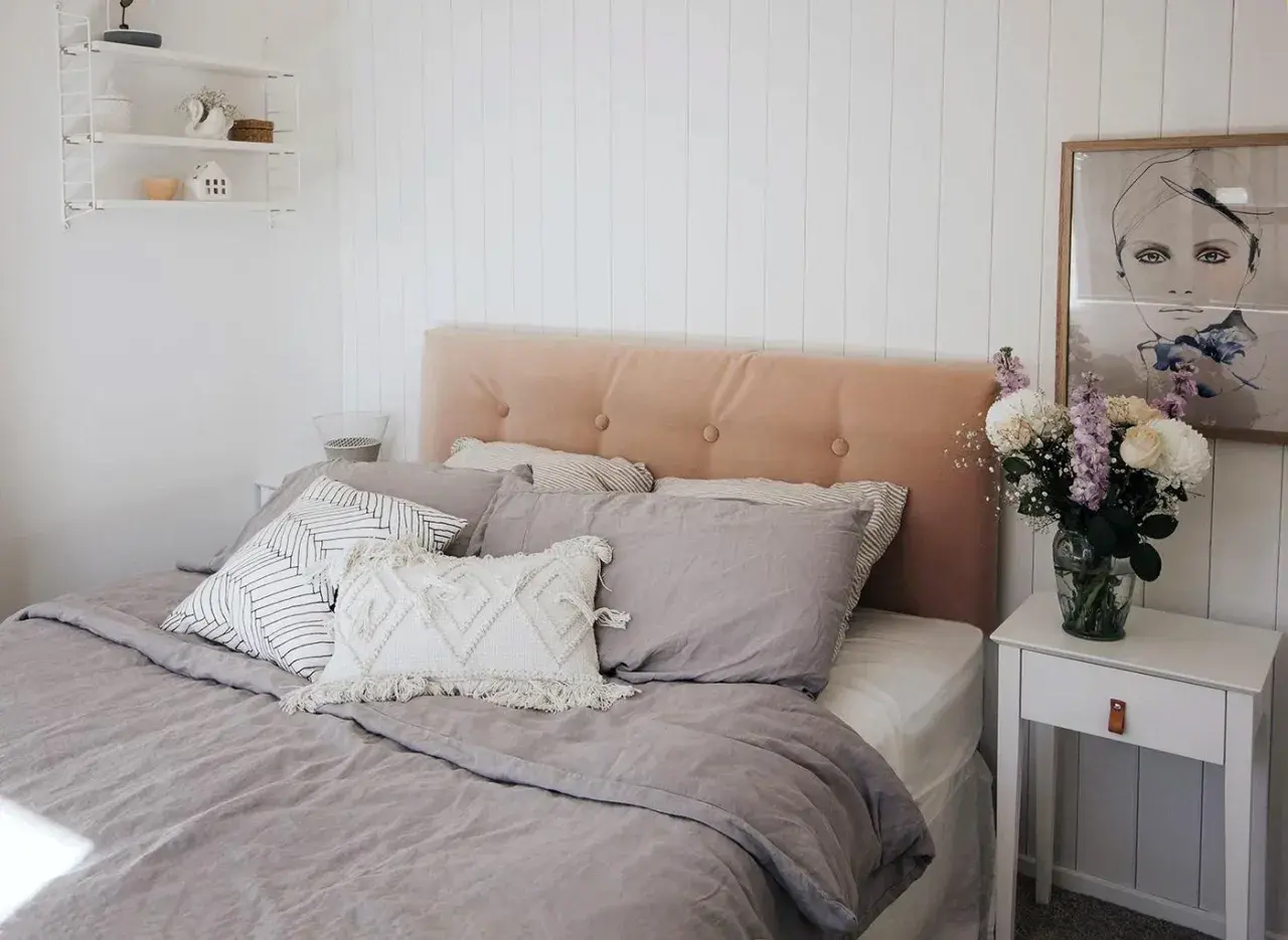 A cozy bedroom featuring a DIY upholstered headboard in a soft peach velvet, adorned with tufted buttons. The bed is dressed in linen bedding and an assortment of decorative pillows.