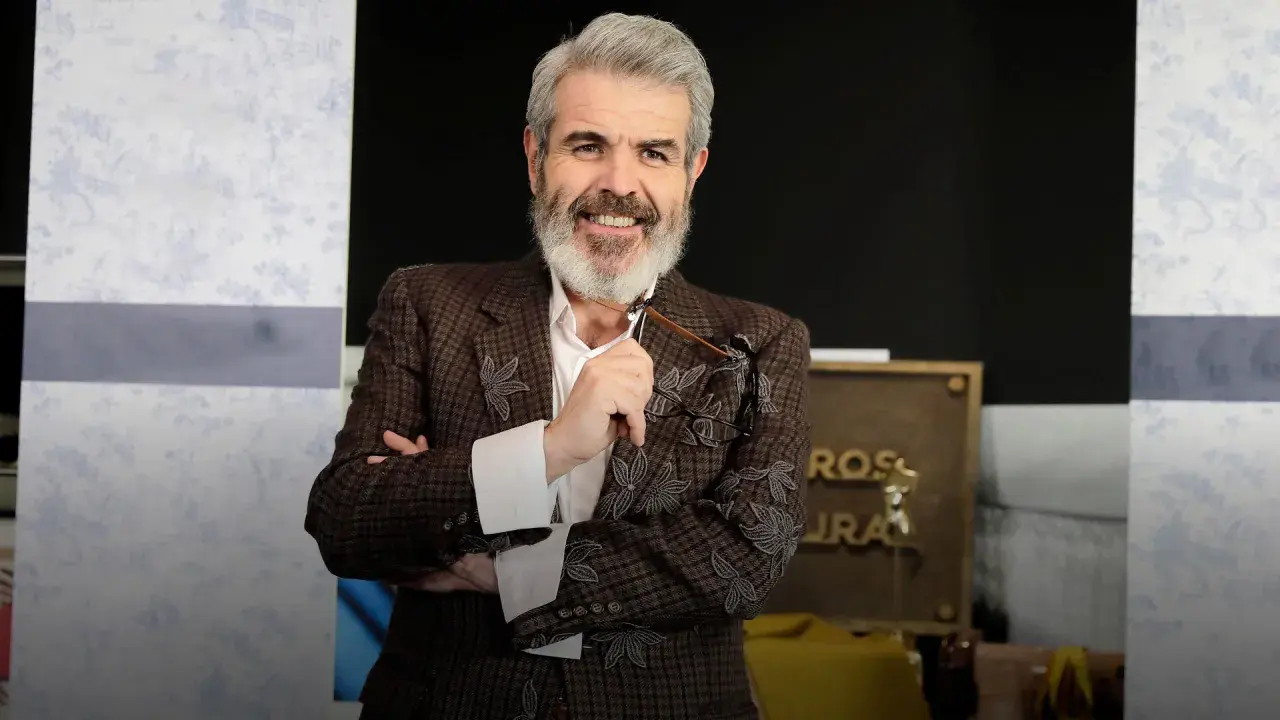 Lorenzo Caprile, con su característica barba canosa, sonríe mientras sostiene unas gafas. Viste una chaqueta de tweed con bordados florales.
