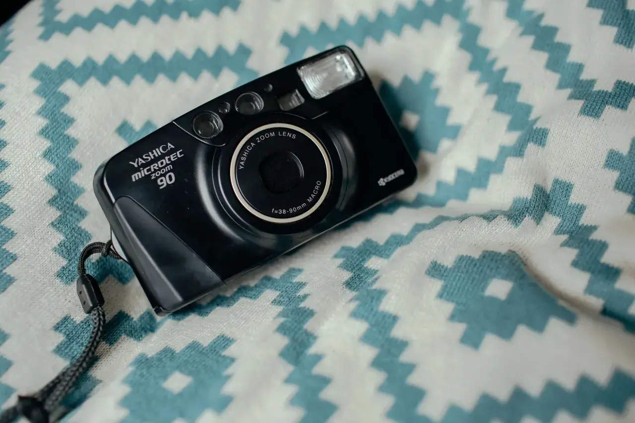 Czarny aparat Yashica Microtec Zoom 90 z paskiem, leżący na białym materiale z turkusowymi zygzakami. Wygląda jak z lat 90.