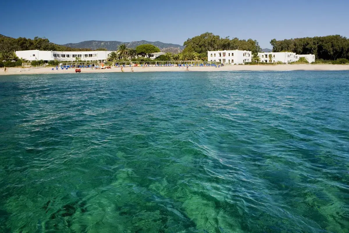 Eaux turquoise cristallines devant une plage de sable fin à Pula, Sardaigne, avec des hôtels blancs et des palmiers.