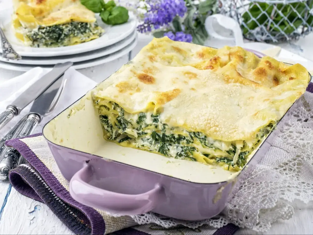 Pyszne lasagne ze szpinakiem w fioletowym naczyniu, gotowe do podania. Obok sztućce i dekoracyjne kwiaty.