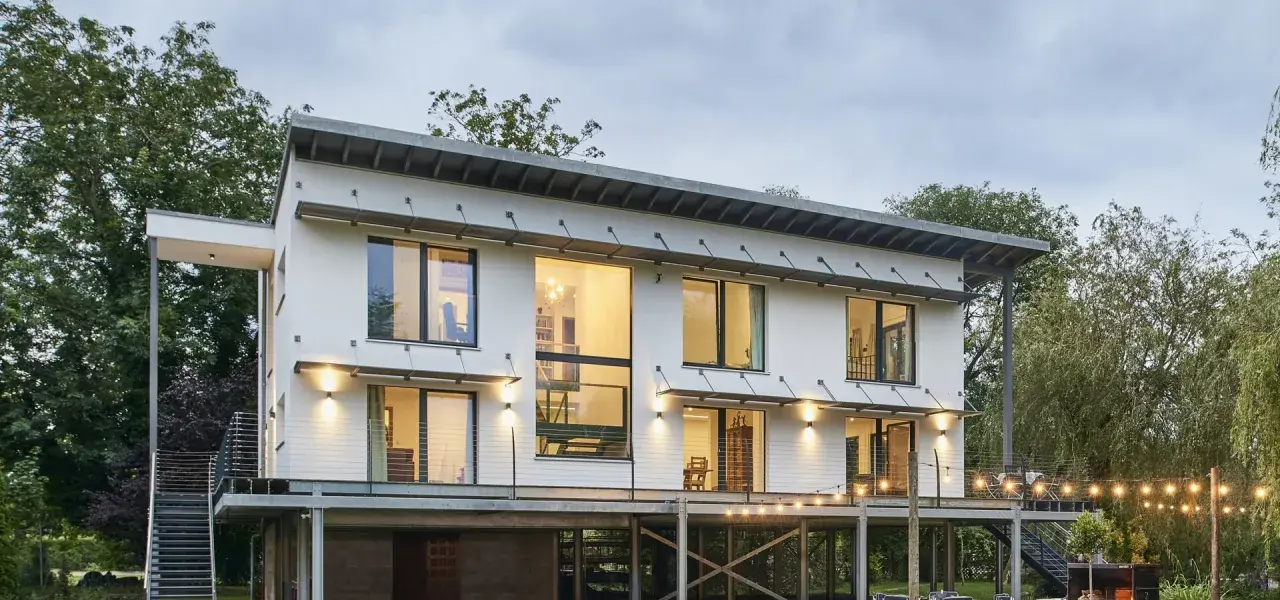 Modernes Haus auf Stelzen mit großen Fenstern und Metalltreppen, umgeben von Bäumen. Lichterketten beleuchten die Terrasse.